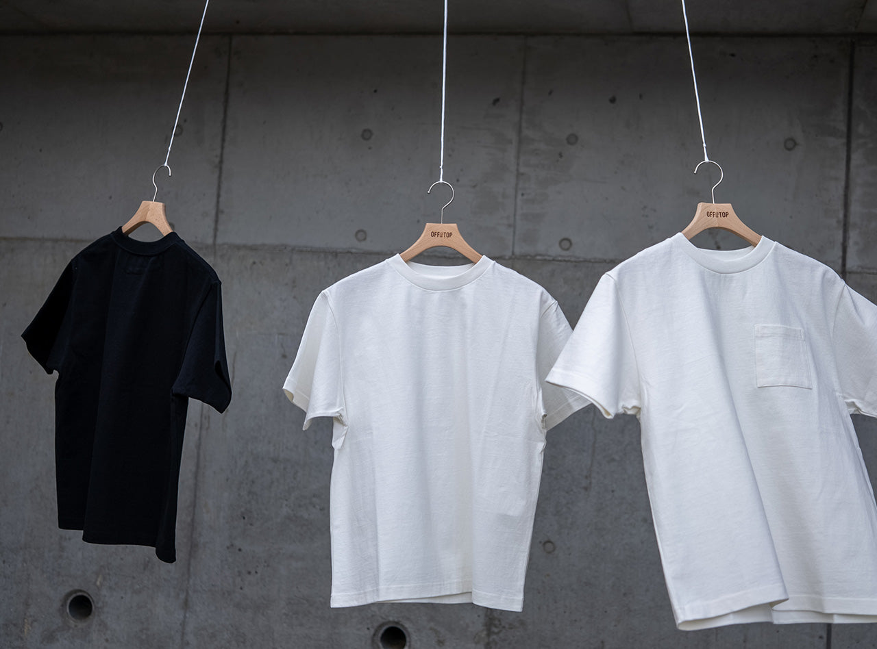 コンクリート壁を背景に吊るされた、10oz（オンス）のヘビーウェイト生地を使用したOFF THE TOPの半袖Tシャツ3型。左からブラック、ホワイト、ホワイト（ポケット付き）。