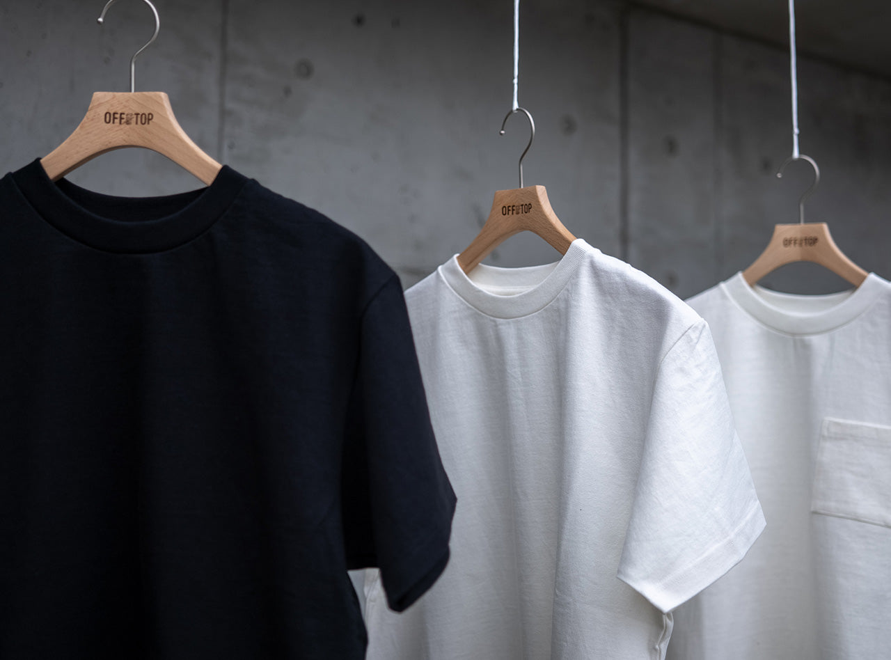 コンクリート壁を背景に吊るされた、10oz（オンス）のヘビーウェイト生地を使用したOFF THE TOPの半袖Tシャツ3型。左からブラック、ホワイト、ホワイト（ポケット付き）。